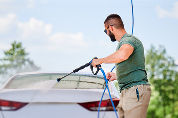 Washing luxury white auto with water gun on an open air car wash. Confident man cleaning his car with high pressure water jet. Technical service concept.