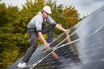 Installer mounting photovoltaic solar panels on roof of house. Man engineer in helmet installing solar modules system outdoors. Concept of alternative and renewable energy.