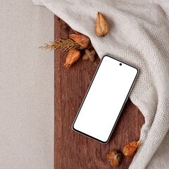 Blank smartphone screen mock up, dried orange flowers on brown wooden board and neutral beige knitted fabric background. Autumn, fall square social media blog, business brand template.