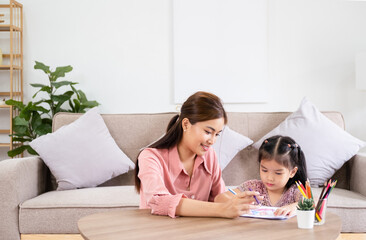 Happy asian girl painting with crayon and colored pencil with his mother in living room at home. Mom teaching daughter how to painting with crayon color on book or doing homework. family concept.