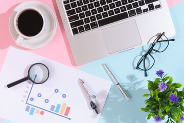 Flowering plant and coffee cup on office desk with laptop, pink and blue background
