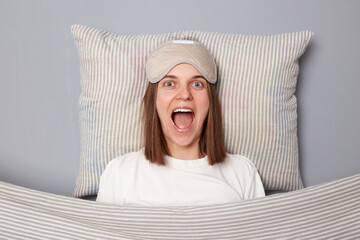 Excited overjoyed woman in white T-shirt and sleeping eye mask lie in bed on pillow under blanket...