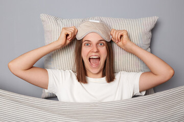 Overjoyed surprised woman in white T-shirt and sleeping eye mask lie in bed on pillow under blanket...