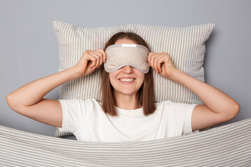 Smiling woman in white T-shirt and sleeping eye mask lie in bed on pillow under blanket isolated on...