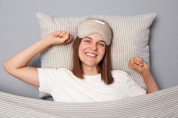 Cheerful happy smiling woman in white T-shirt and sleeping eye mask lie in bed on pillow under...