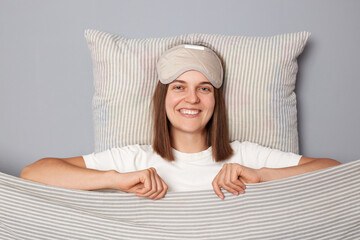 Joyful cute woman in white T-shirt and sleeping eye mask lie in bed on pillow under blanket...