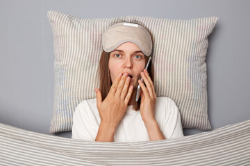 Surprised woman in white T-shirt and sleeping eye mask lie in bed on pillow under blanket isolated...