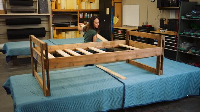 A Young Woman Smiles Enjoying Herself as she Builds an Entire Bed Frame One Piece of Oak Wood at a Time in a Professional Carpentry Workshop.
