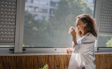 Young smiling caucasian woman in a white shirt standing on the balcony in the morning drinking water and meeting a new day