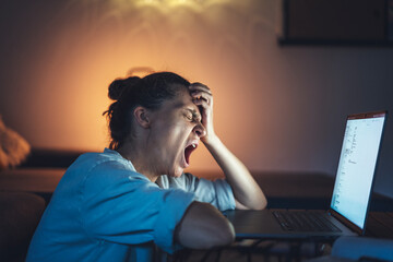 Young woman yawning in front of laptop screen while working from home at night. Deadline and...