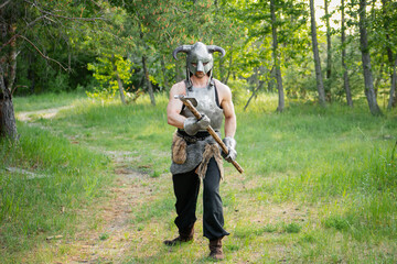 Fototapeta premium Portrait of a warrior in a horned helmet, a steel breastplate with a two-handed ax in his hands, posing against the backdrop of the forest. 