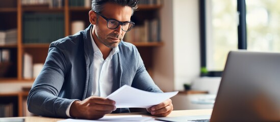 Concentrated businessman from Hispanic background working in office using laptop for online market research while holding documents