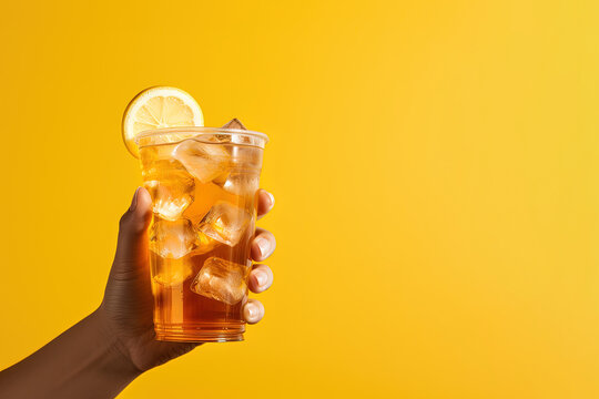 A hand holding takeaway plastic cup of delicious iced lemon tea isolated on yellow background with copy space