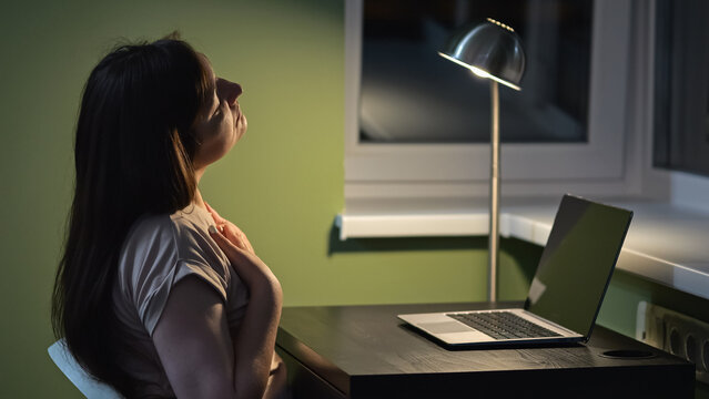 Woman kneads painful neck after working at laptop for long time in premise with floor lamp. Concept of fatigue and overworking. Online profession at home