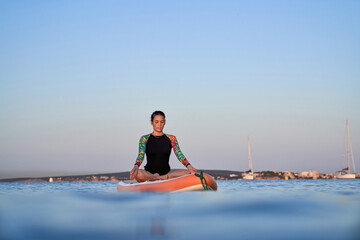 Concentrated ethnic woman meditating in Padmasana on paddleboard