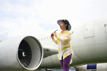 Happy 60s woman in summer clothing and sunglasses walking on runway near airplane. Travel and summer vacation concept