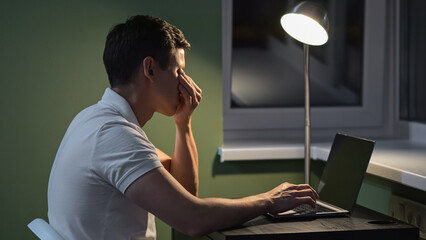 Man feels pain in eyes from load of working at laptop in semi-dark premise with floor lamp. Male...