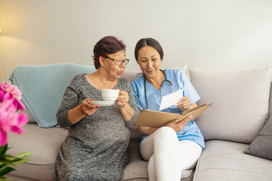 Young doctor visiting elderly woman at home. Happy grandma in glasses holding cup of coffee and watching album with photos with her nurse. Nurse and elderly women in light room