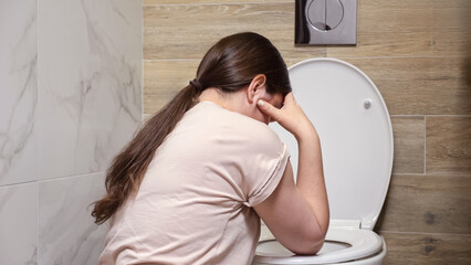 Woman feels nauseous sitting near toilet in bathroom. Feeling unwell after spending time at party. Consequences of drinking alcohol backside view