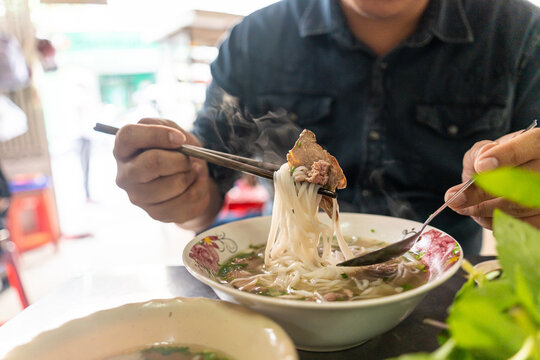 A man using chopsticks and spoon eating traditional Pho Bo vietnamese soup with beef and rice noodles on a metal table, real scene in local restaurant