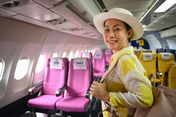 Happy 60s female tourist with bag walking the aisle on plane. Retirement, travel and summer...