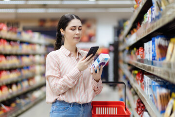 Portrait of smiling Caucasian woman scan qr code of product using smartphone. Shelves with food in...