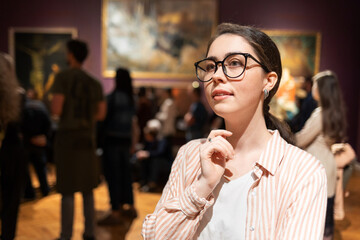 Portrait of young Caucasian woman wearing glasses and looking at exhibition. Masterpieces in...