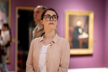 Portrait of young Caucasian woman wearing glasses and looking at exhibition. Masterpieces in background. Cultural education. Concept of Museum Day