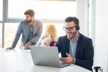 Smiling businessman with headset working on laptop, talking to client, answering questions at office.