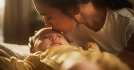 Portrait of Affectionate Asian Mother Leaning Down to Kiss her Baby who is Resting on a Bed. Woman Expressing her Motherly Love to her Infant, Thankful for the Blessing of Having Beautiful Children. - obrazy, fototapety, plakaty