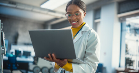 Portrait of Young Black Female Specialist in a Lab Coat Smiling and Using Laptop Computer. Professional and Successful Woman Working as an Engineer, Developing High Technology Projects