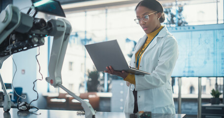 Naklejka premium Portrait of Young Black Female Specialist in Lab Coat Using Laptop Computer to Test an AI Robotic Prototype. Professional, Successful Woman Working as an Engineer in High Tech Company Startup