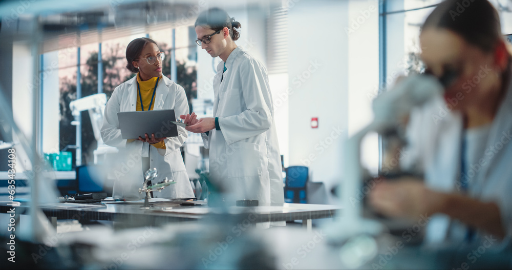 Poster Diverse Young Group of Engineers Working in a Startup Lab. Female Specialist Checking on Electronic Circuit Using Microscope While Two Project Managers Discuss Manufacturing Using Laptop Computer