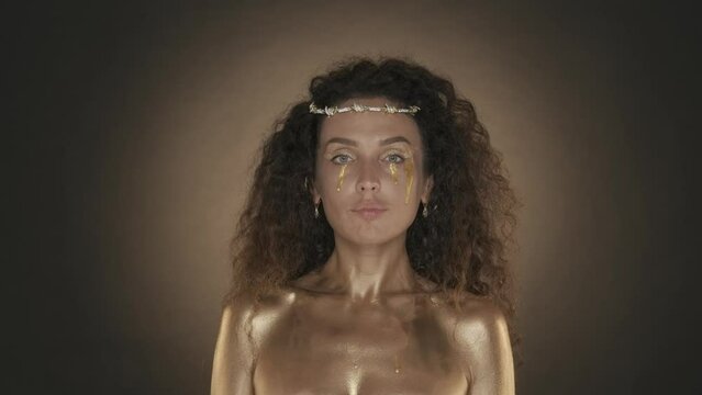 Attractive curly haired young woman with golden makeup smiling and looking at the camera, golden paint and accessories, crown of thornes on the head, close up video. HDR BT2020 HLG Material.