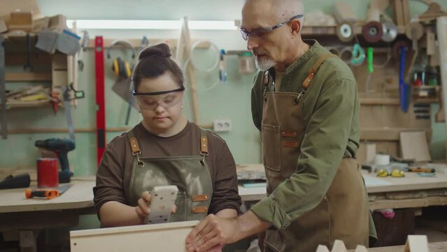 Tilt up of senior craftsman in workwear learning his apprentice with Down syndrome how to polish wooden board during process of preparing workpieces for chair restoration in workshop