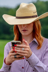 A Lovely Red Headed Cowgirl Uses Her Cellphone In A Country Western Setting Outdoors