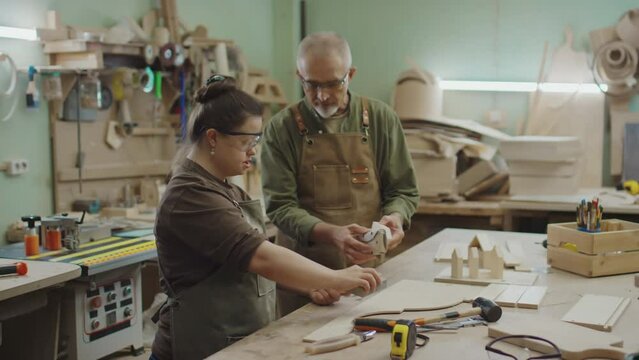 Young woman with Down syndrome and mature male master polishing wooden details with handtools together while standing by workbench with workpieces and and worktools