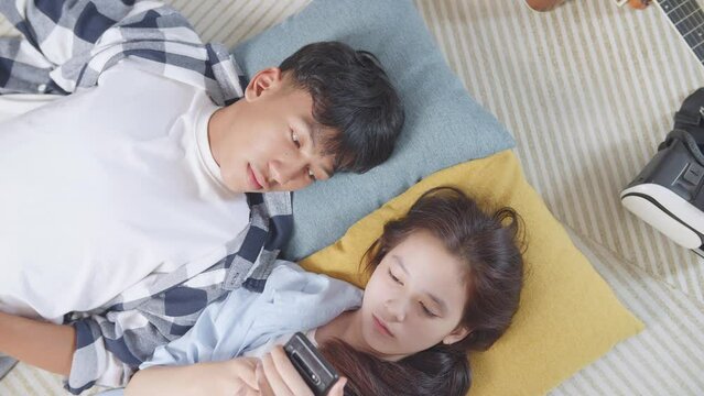 Close Up Top View Of Asian Teen Couple Lying On Carpet On The Floor At Home. Angry Girl Playing Smartphone And Ignoring Her Boyfriend, Upset Boy Leaving His Grilfriend Alone After Trying To Make Her P