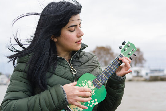 young latin woman traveling outdoors on cold and cloudy day playing ukulele