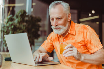 Retired man in summer casual clothes using laptop, watching video drinking lemonade, sitting in cafe