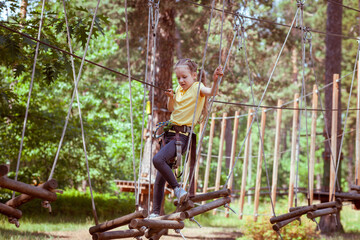 A child in a forest adventure park made of ropes. The girl is climbing the path with a high rope. Children's outdoor climbing entertainment center. Playground for children and sports with a cable car.