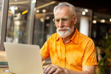 Smiling mature freelancer wearing glasses wearing stylish orange shirt, using laptop, working online