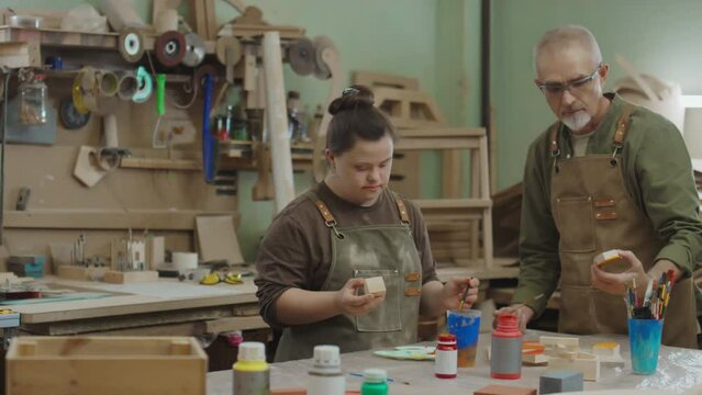 Experienced joiner passing small wooden cube to his apprentice with Down syndrome and teaching her how to paint details while both standing by workbench with many plastic bottles containing gouache