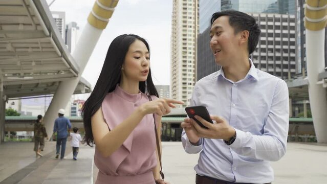 Young Asian business people using a mobile phone and discussing while walking near office buildings. Attractive Asia business woman and man talking - 4K High Quality Footage