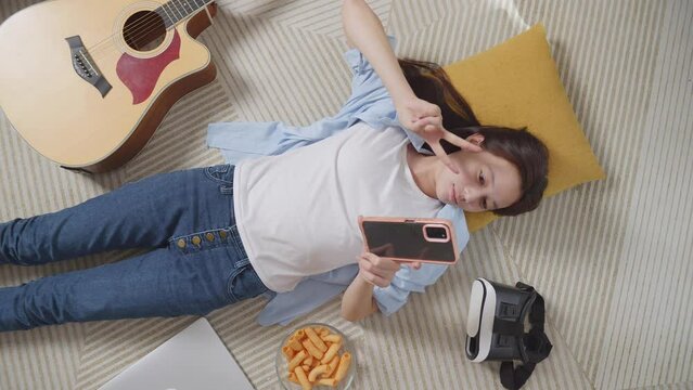 Top View Of Asian Teen Girl Using Smartphone Taking Selfie While Lying On Carpet On The Floor At Home. Smiling, Showing Peace And Heart Gesture
