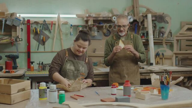 Mature male carpenter and his female apprentice with Down syndrome unwrapping appetizing homemade sandwiches and biting them while standing by workplace and having lunch break