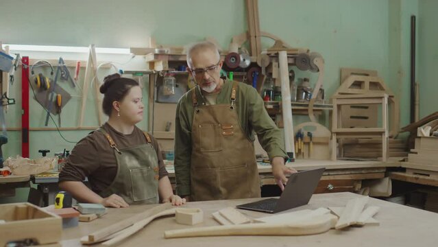 Professional woodworker in apron explaining information about details on laptop screen to his apprentice with Down syndrome and showing her some workpieces on table in workshop