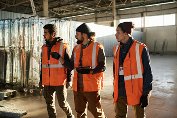 Three young intercultural builders in safety jackets walking along spacious workshop and discussing...