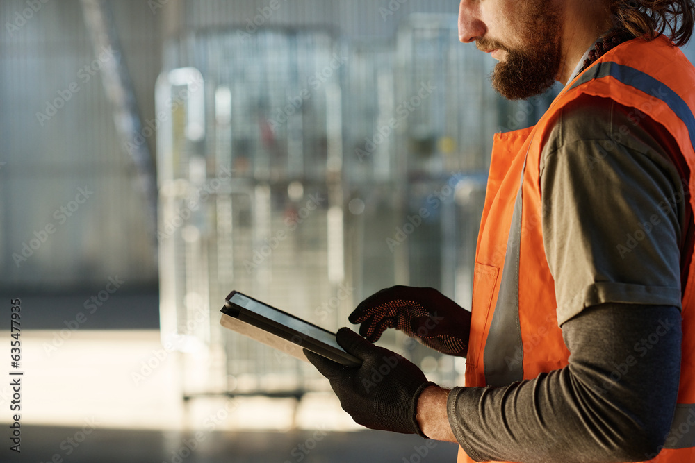 Leinwandbilder side view of young bearded engineer in protective gloves and reflective vest scrolling through sketc