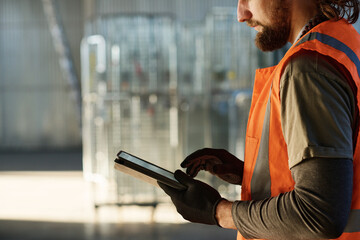Side view of young bearded engineer in protective gloves and reflective vest scrolling through sketch on tablet screen with copyspace on left
